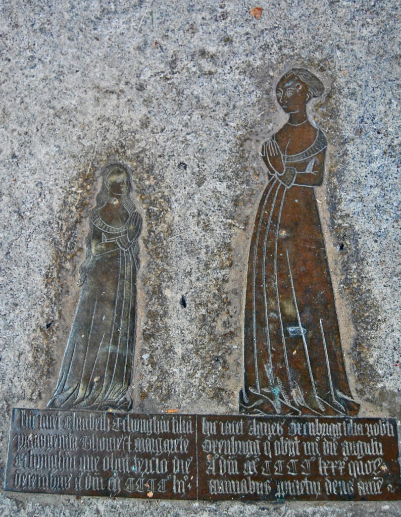 A memorial brass of two women standing side by side looking into each other's eyes. It is mounted on a grey stone wall. The woman on the left is smaller with her hair down. The woman on the right taller with her hair up. Both wear long gowns that brush the ground. The inscription is underneath the figures. 