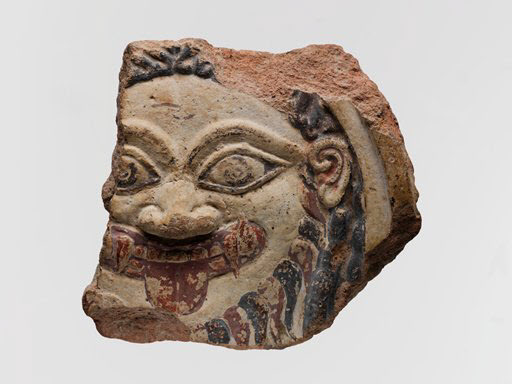 A fragment of a relief sculpture painted with burgundy and black of the head of Medusa with two curved tusk and a round u-shaped tongue protruding from her/their mouth, a wavy beard, a rounded face, and dark hair.